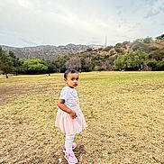 Madina joined the competition — help win amazing prizes! toddler, child, girl, pink_dress, white_tshirt, white_leggings, pink_shoes, grass, field, hill, trees, hollywood_sign, outdoor, sky, cloudy, standing, serious_expression, nature, daytime, park