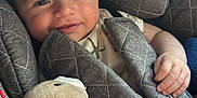 James is registered to the contest to win money with this photo: baby, child, car_seat, quilted, blanket, teddy_bear, soft_toy, smiling, happy, infant, face, hand, comfort, cozy, seat_belt, clothing, portrait, indoors, natural_light, cute