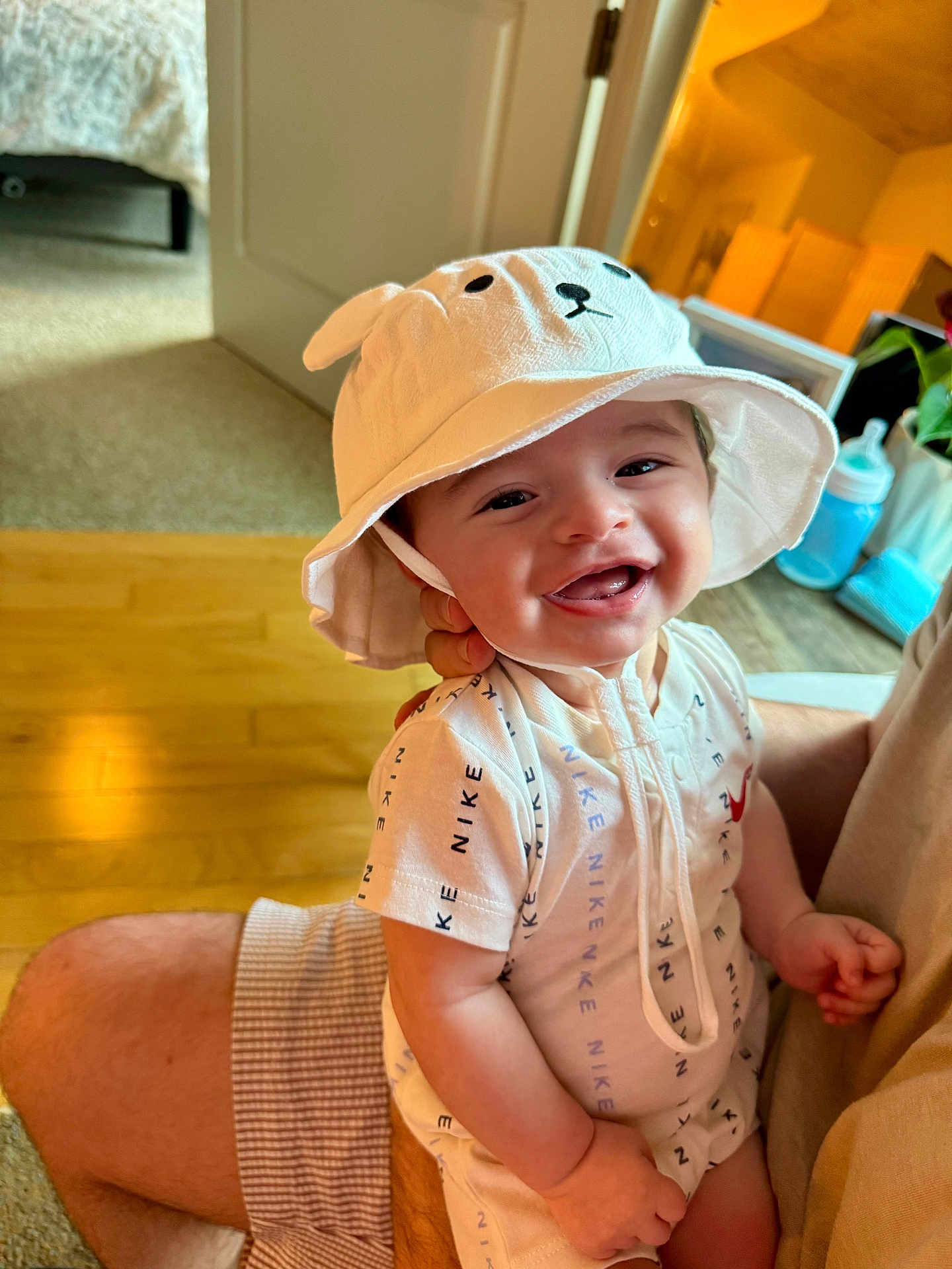 James is registered to the contest to win money with this photo: baby, child, smiling, hat, white_hat, onesie, nike, indoor, carpet, hardwood_floor, baby_bottle, person, holding, happy, cute, face, hand, adult, clothing, infant