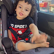 Abdul is registered to the contest to win money with this photo: child, toddler, car_seat, curly_hair, black_shirt, spiderman, waving_hand, car_interior, seatbelt, window, daylight, cute, smiling, barefoot, shorts, seat, kid, young_child, casual_clothing, happy