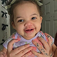 Alaya is registered to the contest to win money with this photo: toddler, child, smiling, tattoos, hands, hair_buns, ribbons, indoor, door, decorations, black_bows, face, person, purple_shirt, floral_pattern, cute, happy, holding, portrait, baby