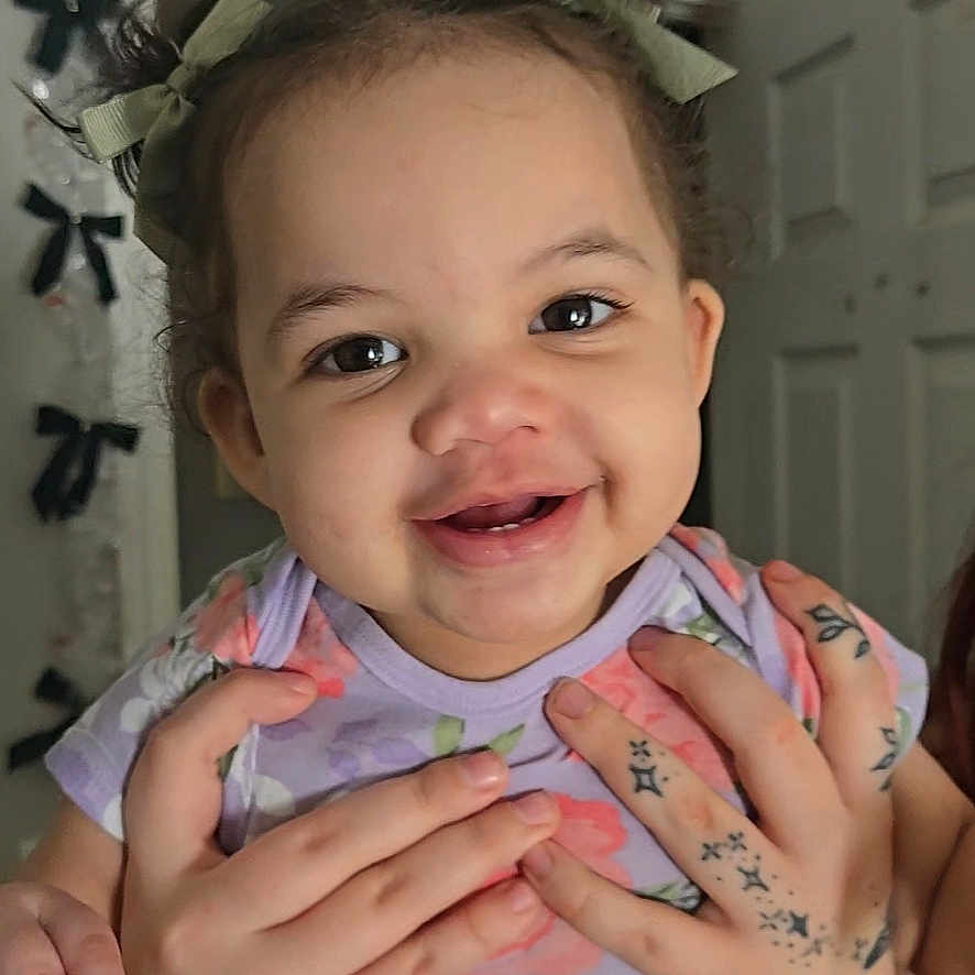 Alaya is registered to the contest to win money with this photo: toddler, child, smiling, tattoos, hands, hair_buns, ribbons, indoor, door, decorations, black_bows, face, person, purple_shirt, floral_pattern, cute, happy, holding, portrait, baby