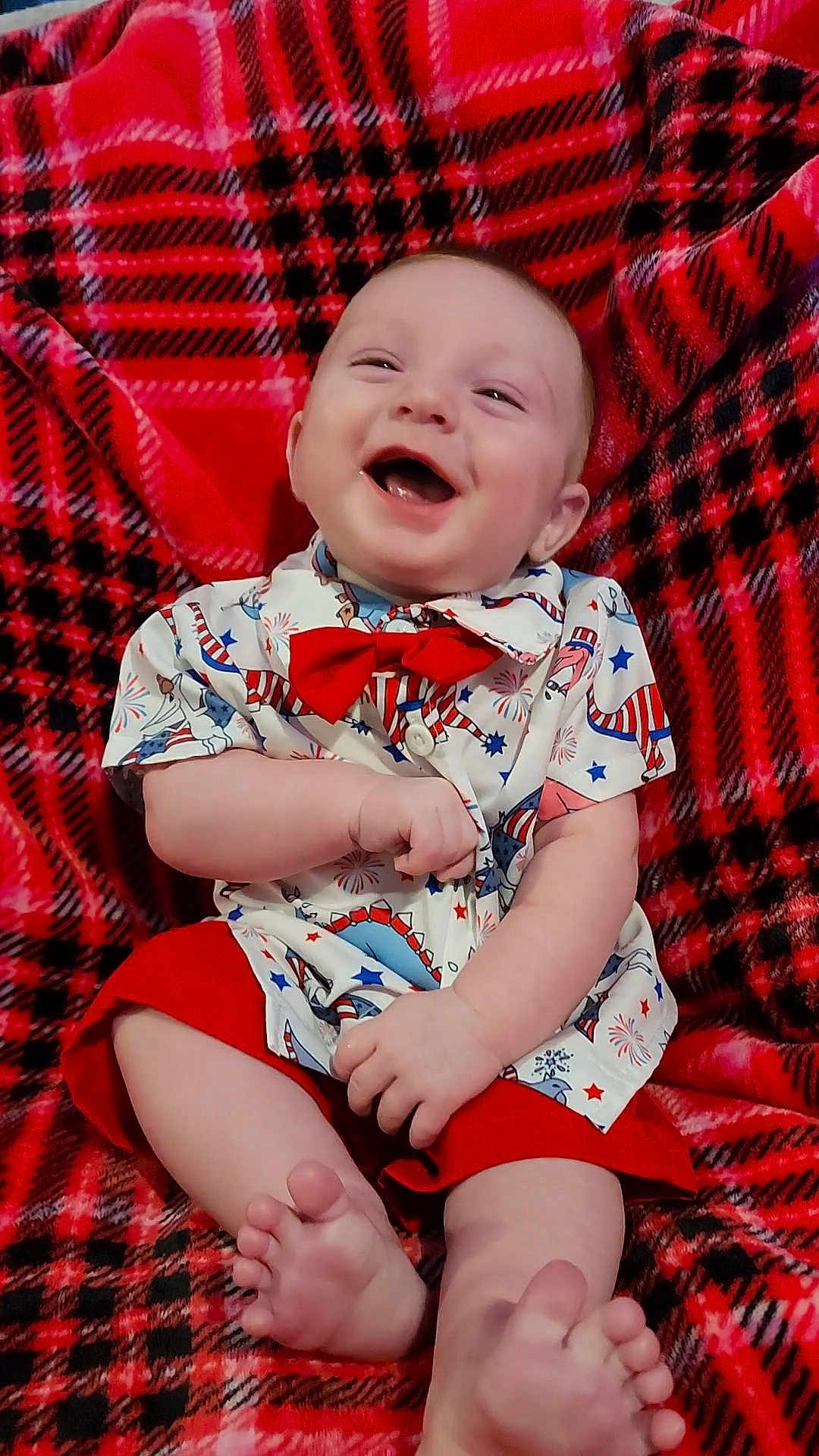 Kylan is registered to the contest to win money with this photo: baby, child, smiling, laughing, red_bow_tie, shirt, shorts, plaid_blanket, feet, happy, cute, indoor, sitting, infant, person, clothing, joyful, portrait, expression, casual