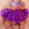 baby, child, infant, portrait, lying_down, purple_dress, tutu, headband, floral_pattern, smiling, wide_eyes, hands, toy, pink_blanket, soft_texture, indoor, colorful, fashion, cute, full_body