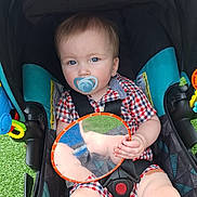 Finn joined the competition — help win amazing prizes! toddler, child, stroller, pacifier, blue_eyes, checkered_clothing, toy_mirror, reflection, sitting, outdoor, green_grass, colorful_toys, baby, infant, seatbelt, black_straps, plaid, cute, face, hand