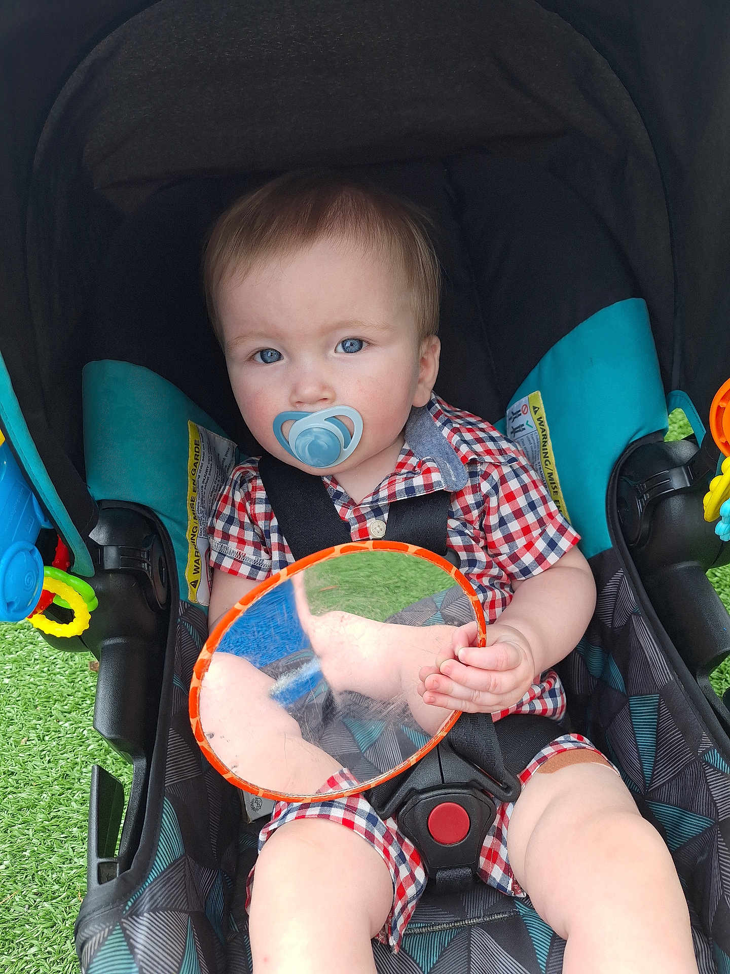 Finn joined the competition — help win amazing prizes! toddler, child, stroller, pacifier, blue_eyes, checkered_clothing, toy_mirror, reflection, sitting, outdoor, green_grass, colorful_toys, baby, infant, seatbelt, black_straps, plaid, cute, face, hand