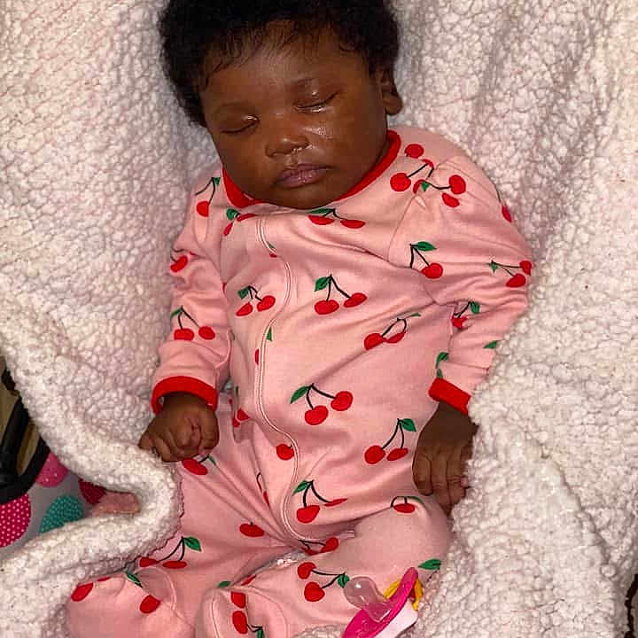 Sarya is registered to the contest to win money with this photo: baby, infant, sleeping, portrait, pink_outfit, cherry_pattern, onesie, blanket, white_blanket, pacifier, curly_hair, dark_skin, closed_eyes, peaceful, indoors, cozy, nursery, lying_down, child, photography