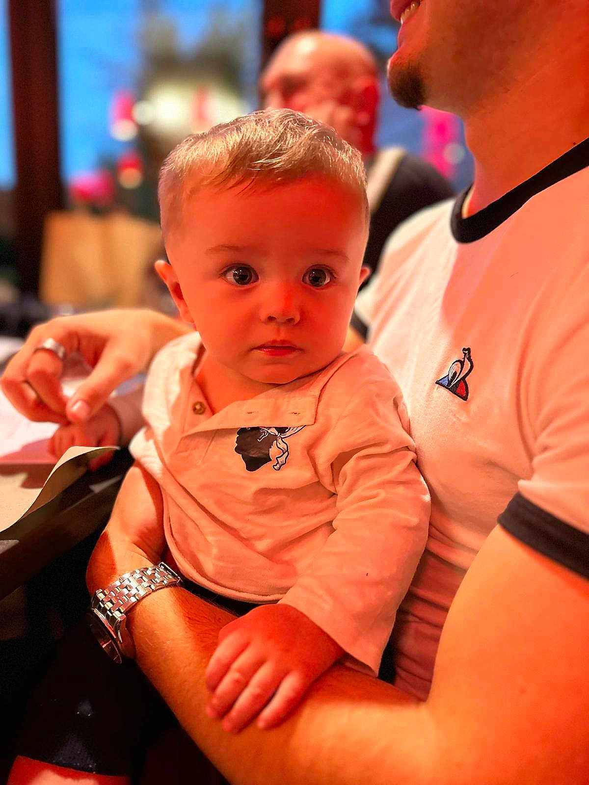 Ezekiel a rejoint le concours — aidez-le/la à gagner de superbes lots ! toddler, child, person, adult, indoor, arm, watch, shirt, curious, face, blond_hair, short_hair, seated, blurred_background, warm_lighting, portrait, closeup, casual_clothing, holding, expression