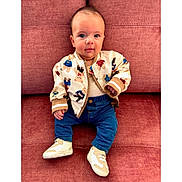 Denver is registered to the contest to win money with this photo: baby, child, jacket, teddy_bear, pattern, jeans, sneakers, couch, red_couch, seated, portrait, cute, indoor, clothing, smile, face, person, young, fashion, casual