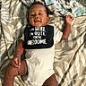 baby, infant, onesie, bib, bed, blanket, striped_socks, smiling, happy, lying_down, child, cute, indoors, person, text_on_bib, patterned_bedspread, skin, face, hand, foot