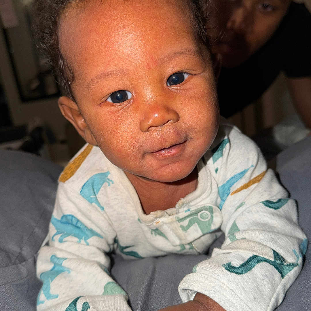Khylo joined the competition — help win amazing prizes! adult, baby, bed, blanket, child, cozy, curious, cute, dinosaur_onesie, face, family, hands, home, indoor, infant, parent, person, portrait, smiling, young_child