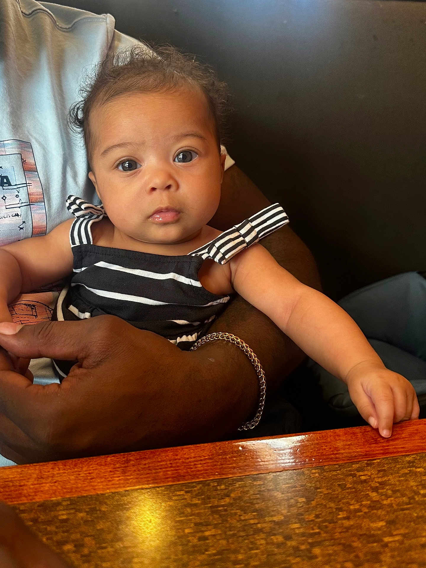 Evelynn joined the competition — help win amazing prizes! baby, child, striped_clothing, person, arm, hand, bracelet, table, wood, indoor, face, curly_hair, eyes, skin, sitting, holding, black_clothing, casual, portrait, cute