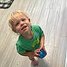 child, toddler, blonde_hair, green_shirt, toy, blue_toy, barefoot, floor, wood_floor, indoor, smiling, looking_up, curious, home, welcome_mat, light, casual_clothing, happy, person, young_child