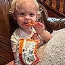 Jackson is registered to the contest to win money with this photo: toddler, child, snack, cheese_puffs, hand, couch, leather, curly_hair, blonde, indoor, stairs, wood, white_shirt, smiling, face, person, food, relaxing, cozy, carpet