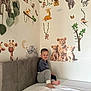 child, bed, wall_stickers, lion, monkey, giraffe, elephant, smiling, indoor, decor, playful, cute, cozy, young_boy, sitting, pajamas, headboard, white_bed_sheet, room, happy