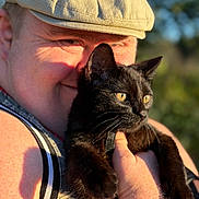 Samwise is registered to the contest to win money with this photo: person, cat, black_cat, yellow_eyes, cap, sleeveless_top, smiling, holding, outdoor, daylight, portrait, closeup, watch, happy, animal, pet, nature, sunlight, face, hand
