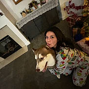 Cacho is registered to the contest to win money with this photo: cabinet, carpet, celebration, christmas_tree, cozy, decorations, dog, festive, fireplace, holiday, home, hug, husky, indoor, pajamas, pet, presents, smile, warm, woman