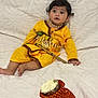 child, baby, yellow_clothing, traditional_dress, necklace, bracelet, decorative_stick, red_pouch, white_blanket, indoor, portrait, cute, sitting, face, curious, soft_texture, cultural, festive, young_child, footwearless