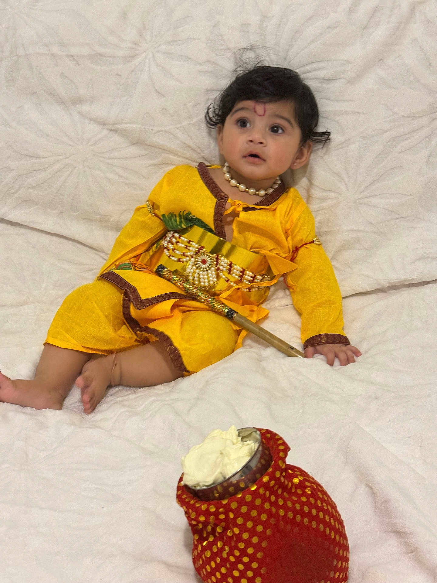 Devansh joined the competition — help win amazing prizes! child, baby, yellow_clothing, traditional_dress, necklace, bracelet, decorative_stick, red_pouch, white_blanket, indoor, portrait, cute, sitting, face, curious, soft_texture, cultural, festive, young_child, footwearless