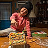 Andrea joined the competition — help win amazing prizes! child, girl, gingerbread_house, cookie, table, pink_sweater, black_hat, smiling, pointing, indoor, wooden_table, home_decor, lamp, curtains, cabinet, cookies, plastic_wrap, happy, cozy, holiday