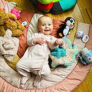 Elaina is registered to the contest to win money with this photo: baby, infant, smiling_baby, play_mat, blanket, stuffed_toy, penguin_plush, crochet_blanket, wooden_floor, onesie, baby_toy, pacifier, soft_toy, plush_bear, happy, lying_down, head, face, play_area, flooring