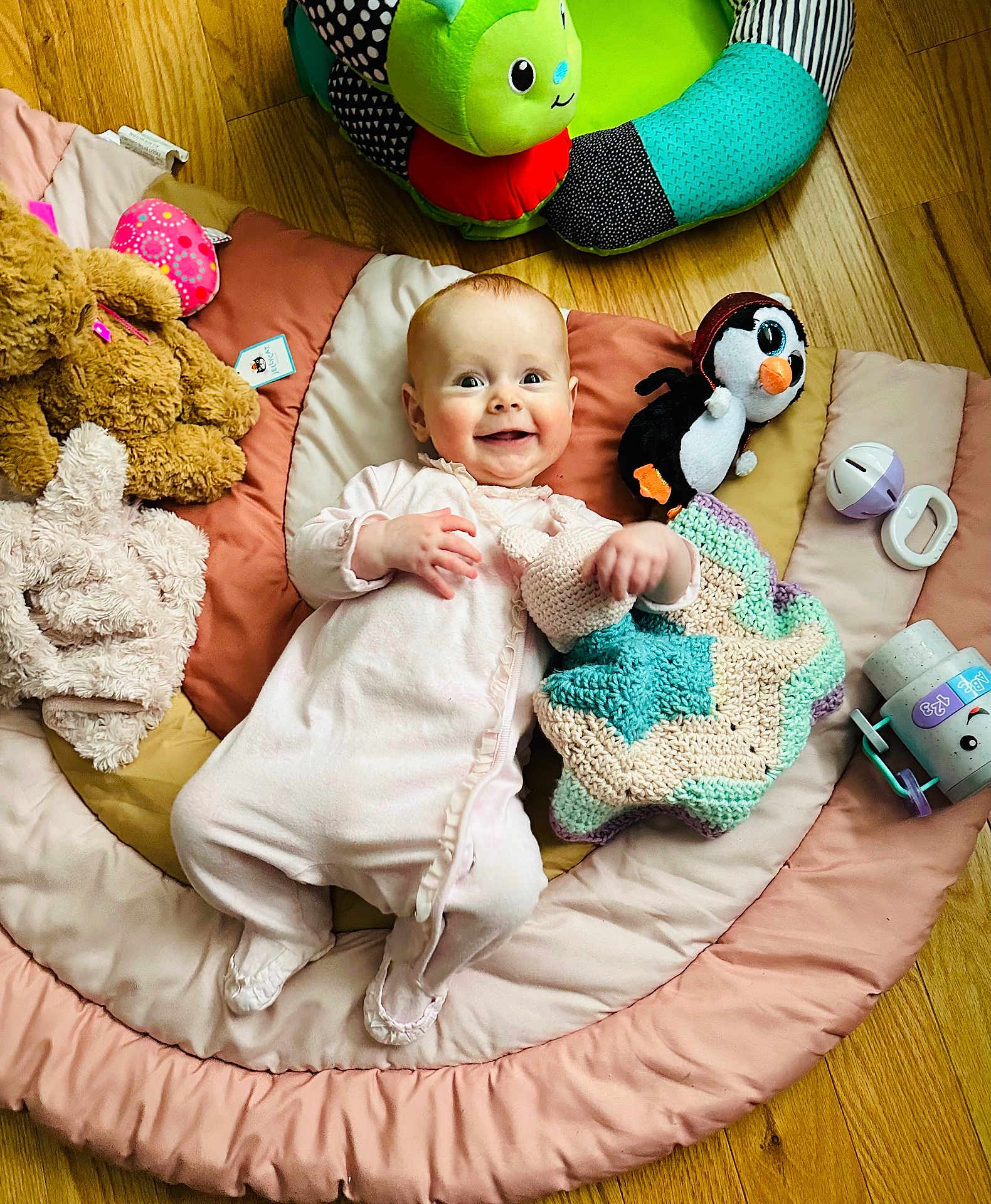 Elaina is registered to the contest to win money with this photo: baby, infant, smiling_baby, play_mat, blanket, stuffed_toy, penguin_plush, crochet_blanket, wooden_floor, onesie, baby_toy, pacifier, soft_toy, plush_bear, happy, lying_down, head, face, play_area, flooring