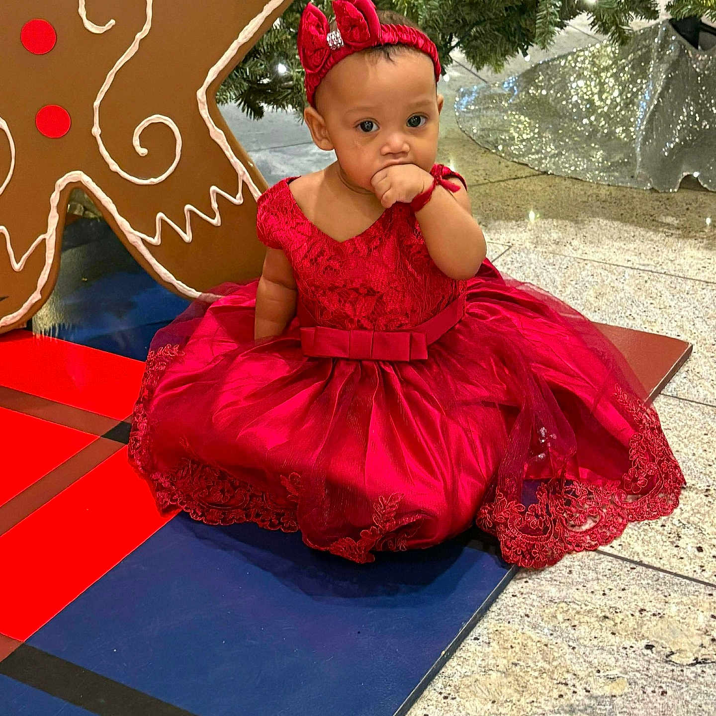 Inara joined the competition — help win amazing prizes! baby, child, red_dress, headband, gingerbread_man, christmas_tree, holiday, decoration, floor, sitting, cute, festive, indoor, portrait, person, holiday_season, sparkle, toy, event, happy