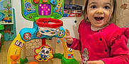 Giuliana a rejoint le concours — aidez-le/la à gagner de superbes lots ! basketball_hoop, carpet, child, colorful, cute, excitement, floor, furniture, game, happy, indoor, orange_ball, person, pink_sweater, playroom, shelf, smiling, toddler, toy, young_child