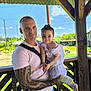 baby, casual, child, cute, daylight, family, flamingos, gazebo, greenery, holding, man, nature, outdoor, people, portrait, smile, sunny, tattoo, white_clothing, wooden_structure