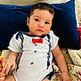 baby, infant, child, onesie, bow_tie, hand_holding, pillow, quilt, floral_pattern, navy_blue, cute, portrait, indoors, skin, face, eyes, sitting, clothing, person, small_hand