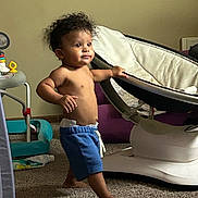 Brashaun joined the competition — help win amazing prizes! toddler, child, baby, curly_hair, blue_shorts, bare_chest, carpet, baby_rocker, toy, indoor, walking, curious, furniture, diaper, foot, hand, face, person, room, home