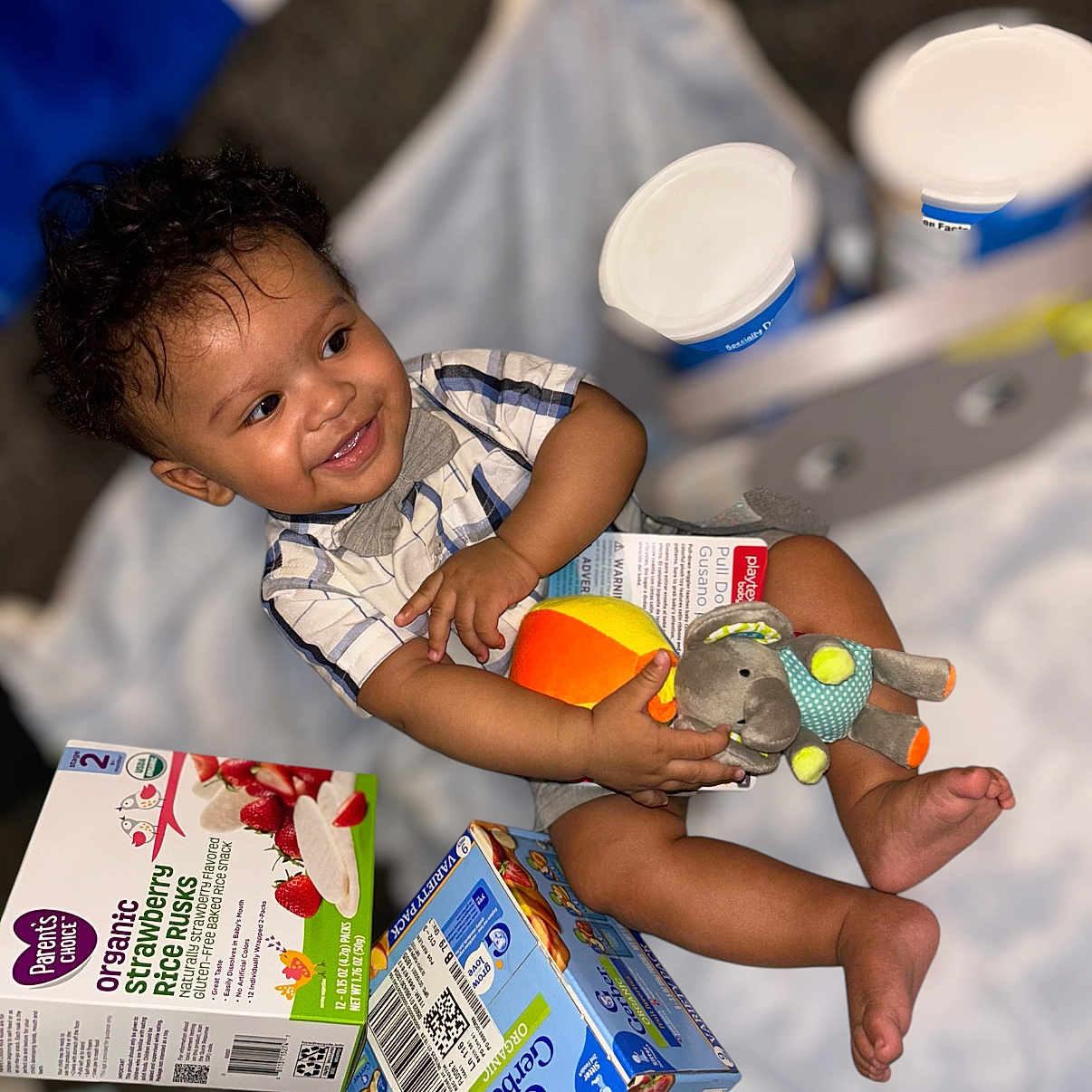 Brashaun is registered to the contest to win money with this photo: baby_clothing, blanket, child, cracker_box, curly_hair, cute, elephant_toy, food_packaging, happy, indoors, infant, orange_toy, organic_snacks, playtime, plush_toy, rice_rusk_box, short_sleeves, sitting, smile, toddler