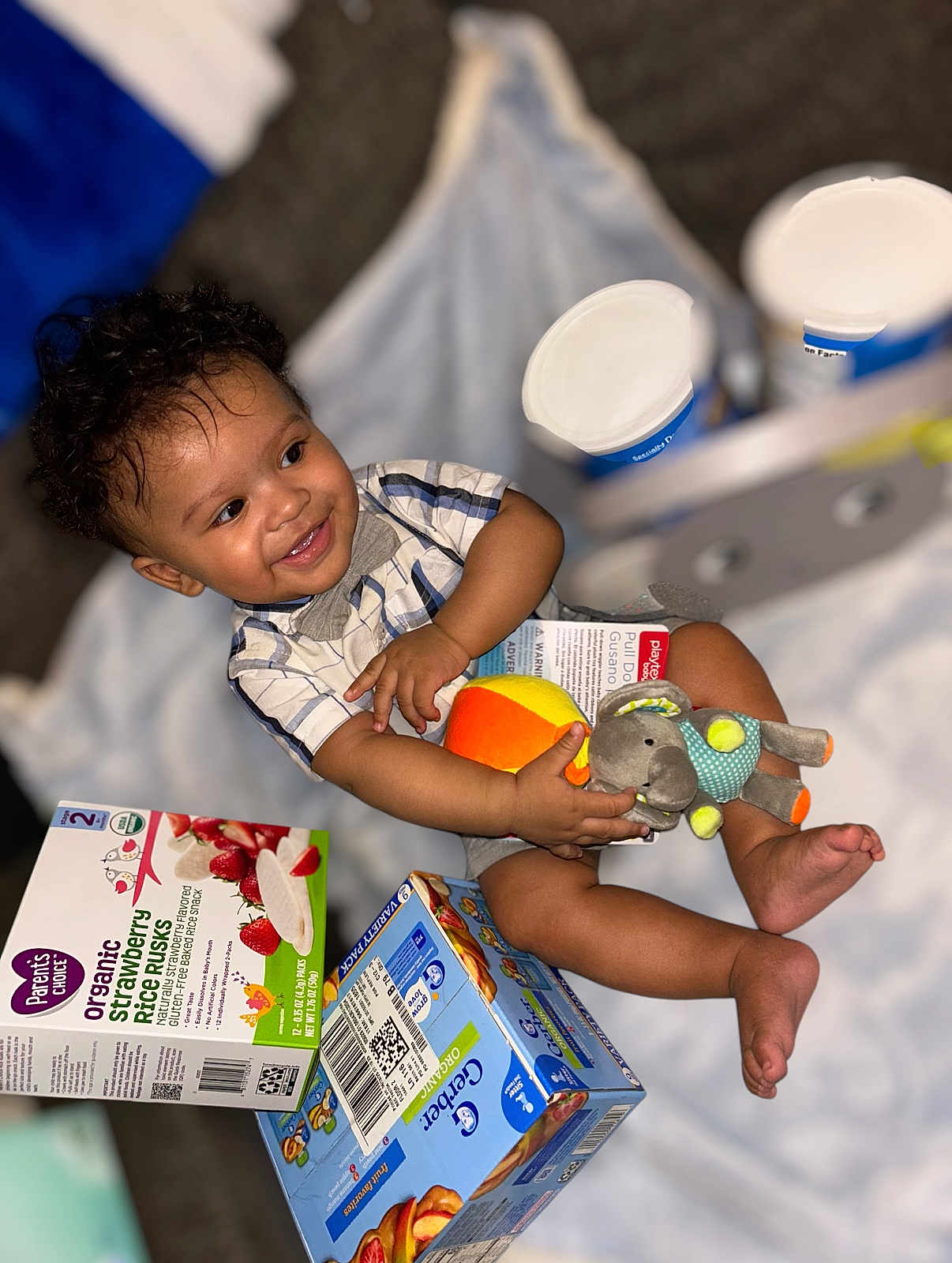 Brashaun is registered to the contest to win money with this photo: toddler, child, smile, curly_hair, plush_toy, elephant_toy, orange_toy, organic_snacks, rice_rusk_box, cracker_box, blanket, sitting, happy, indoors, baby_clothing, short_sleeves, food_packaging, playtime, cute, infant