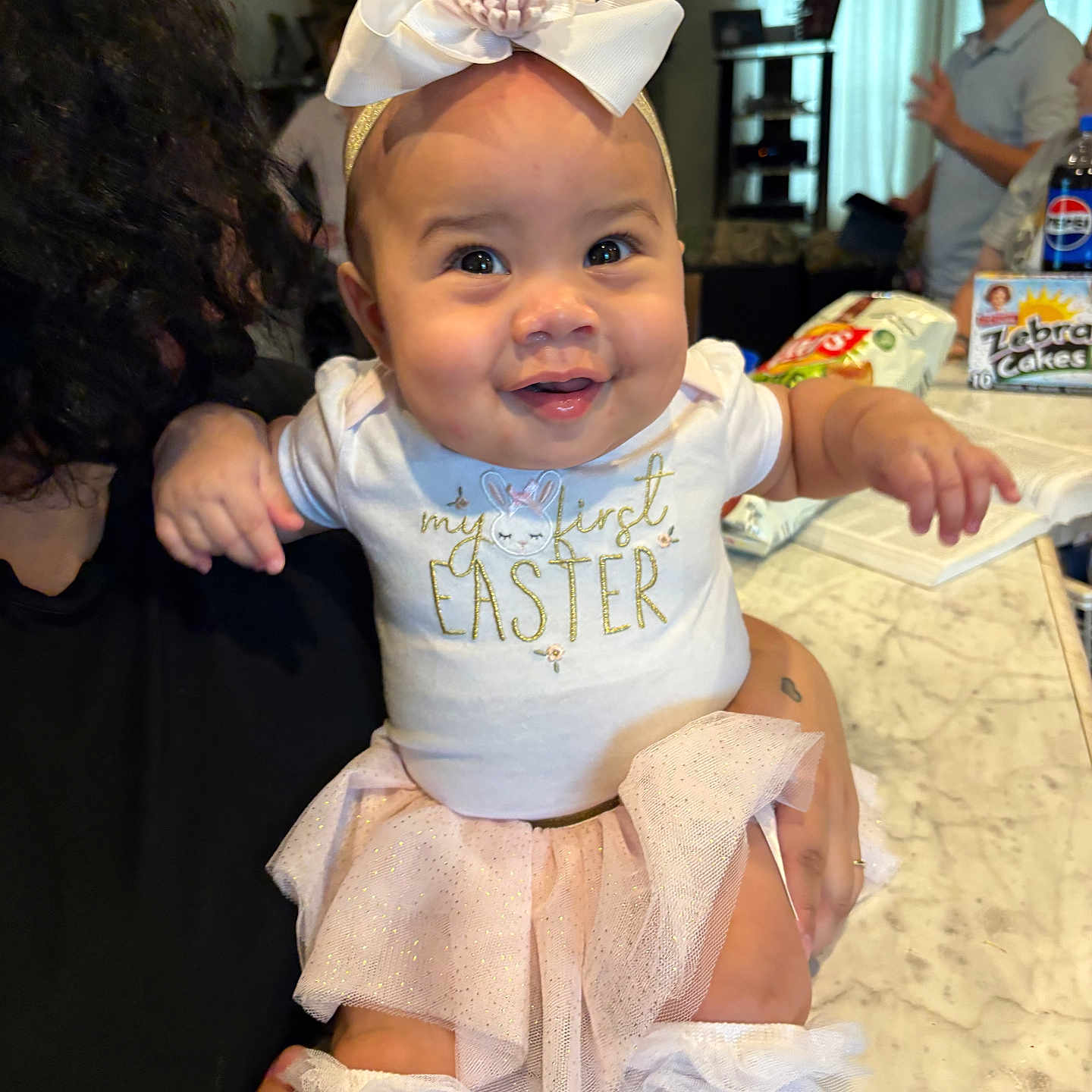 Naomi is registered to the contest to win money with this photo: baby, child, infant, smiling, bow, headband, tutu, white_clothing, kitchen, countertop, person, adult, man, snack, beverage, indoor, celebration, happy, family, portrait
