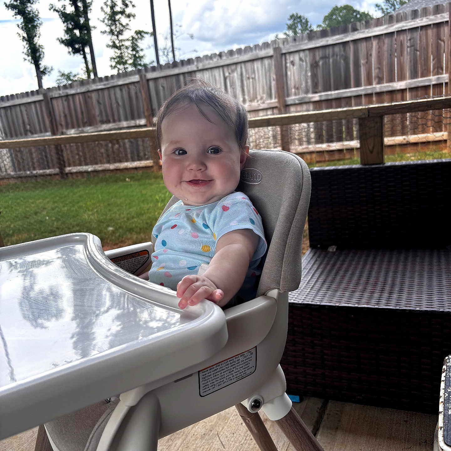 Preslee Rose Ingram is registered to the contest to win money with this photo: baby, child, high_chair, smiling, outdoor, backyard, patio, wooden_fence, grass, trees, sky, clouds, infant, portrait, sitting, blue_clothing, polka_dots, feeding_tray, furniture, daylight
