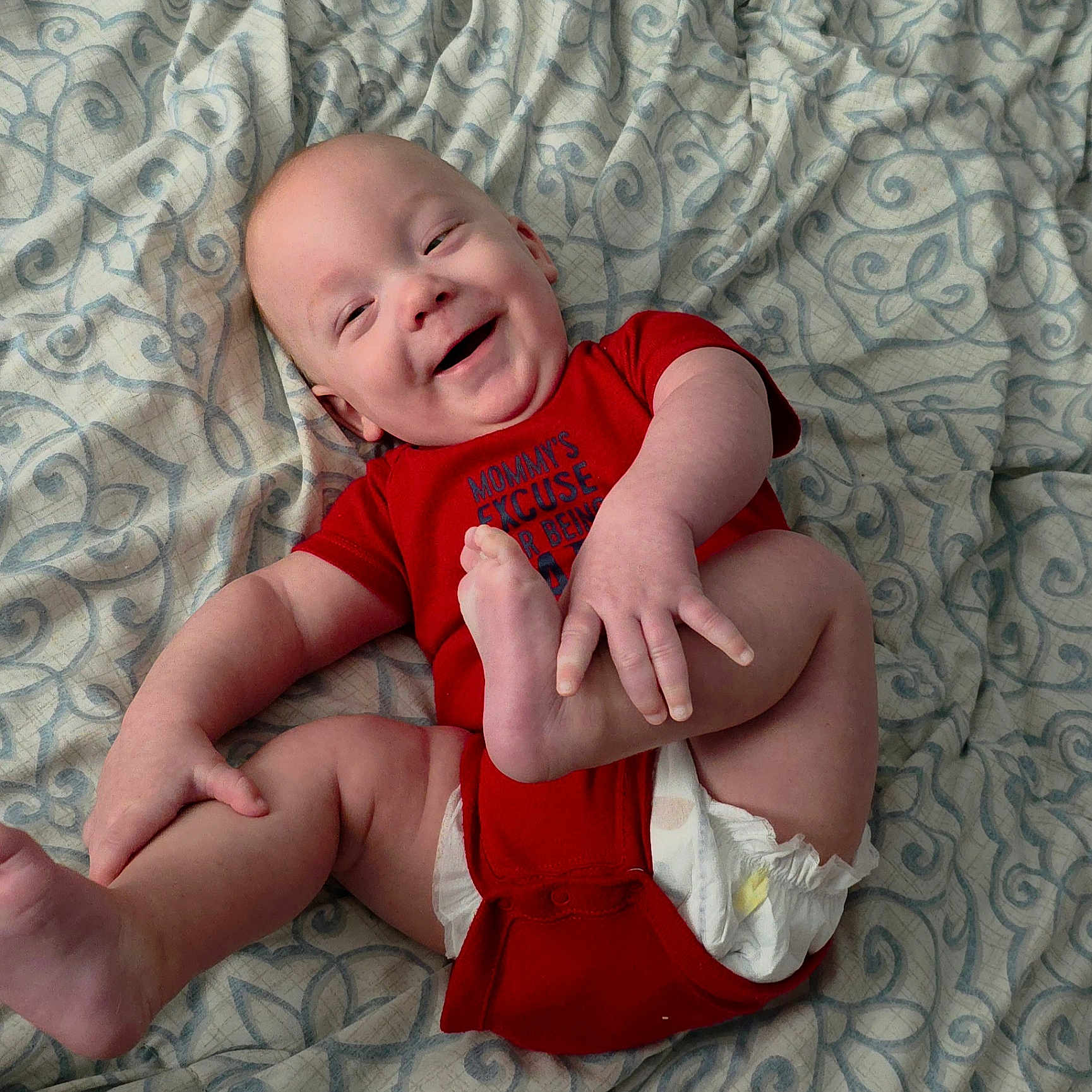 John joined the competition — help win amazing prizes! baby, infant, smiling, red_onesie, diaper, barefoot, lying_down, blanket, patterned_blanket, happy, child, cute, playful, skin, hand, foot, person, indoors, portrait, toddler