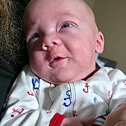 John is registered to the contest to win money with this photo: baby, infant, onesie, clothing, face, hand, expression, cute, indoor, person, child, newborn, portrait, holding, skin, blue, red, white, closeup, smile