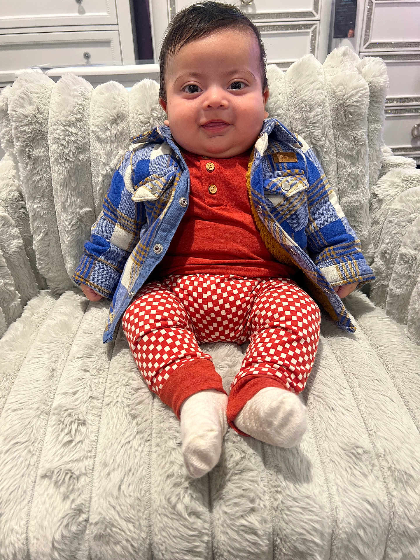 Christopher is registered to the contest to win money with this photo: baby, child, smiling, plaid_jacket, patterned_pants, socks, fluffy_chair, indoor, cute, portrait, seated, happy, cozy, face, furniture, warm_clothing, infant, young_child, person, candid