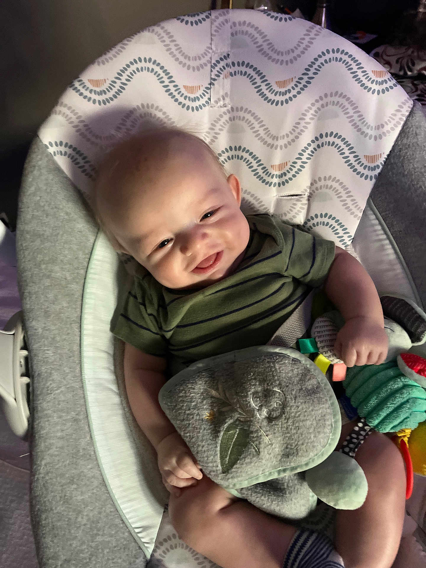 Luca is registered to the contest to win money with this photo: baby, infant, smiling, bouncer, toy, plush_toy, striped_shirt, onesie, face, hands, legs, seat, blanket, patterned_fabric, cozy, indoor, portrait, closeup, happy, lovey