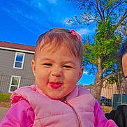 Nashly is registered to the contest to win money with this photo: toddler, child, pink_clothing, puffy_vest, smile, face, outdoor, tree, blue_sky, house, window, fence, grass, person, daylight, playful, cute, hair_bow, expression, portrait