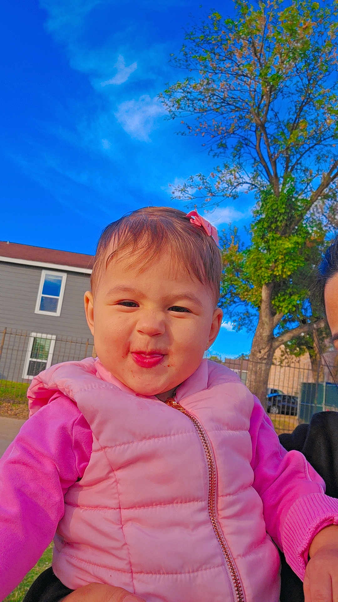 Nashly is registered to the contest to win money with this photo: toddler, child, pink_clothing, puffy_vest, smile, face, outdoor, tree, blue_sky, house, window, fence, grass, person, daylight, playful, cute, hair_bow, expression, portrait