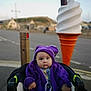baby, child, purple_blanket, purple_hat, stroller, ice_cream_sculpture, outdoor, street, road, sidewalk, blurred_background, daytime, curious_expression, winter_clothing, urban, child_portrait, infant, cozy, wide_eyes, soft_lighting
