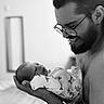 Stetson is registered to the contest to win money with this photo: man, baby, newborn, black_and_white, glasses, beard, holding, smile, indoor, portrait, intimate, family, love, tenderness, clothing, onesie, person, face, head, human
