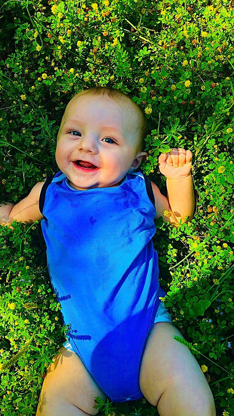 Briar is registered to the contest to win money with this photo: baby_toddler_clothing, blue, botany, eye, face, facial_expression, flower, gesture, grass, green, happy, joy, leaf, light, people_in_nature, person, photograph, plant, skin, smile