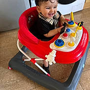 Adem participe au concours pour gagner de l'argent avec cette photo : toddler, baby_walker, child, toy, indoor, floor, curly_hair, smiling, playful, red, gray, kitchen, person, clothing, black_shirt, plastic_toy, happy, walker, footwear, home