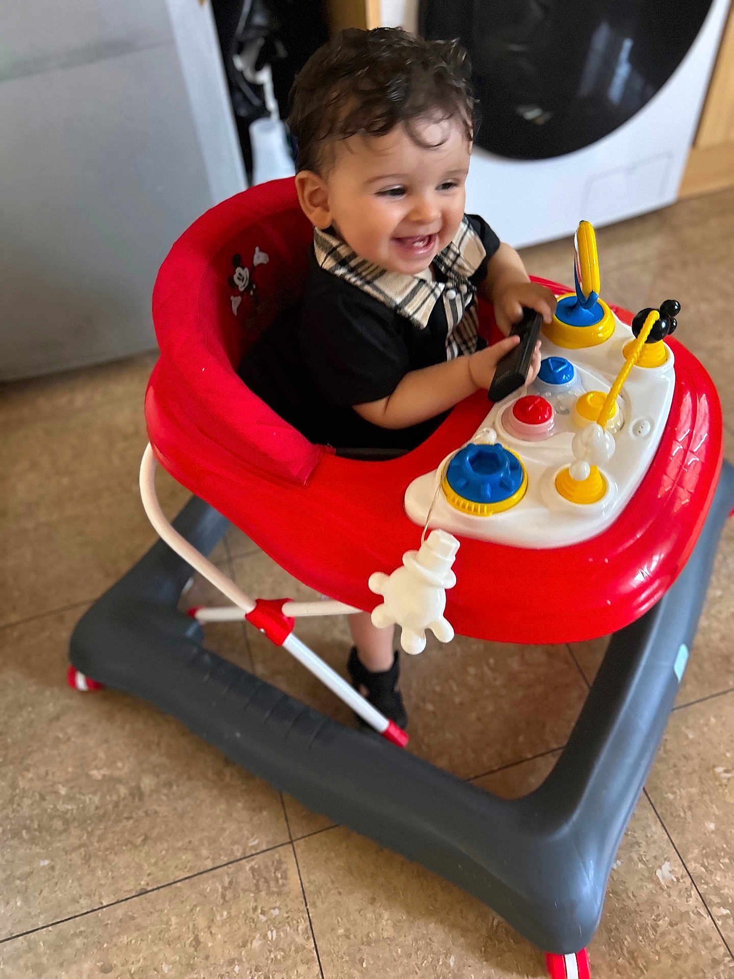 Adem participe au concours pour gagner de l'argent avec cette photo : toddler, baby_walker, child, toy, indoor, floor, curly_hair, smiling, playful, red, gray, kitchen, person, clothing, black_shirt, plastic_toy, happy, walker, footwear, home