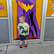 Joshua joined the competition — help win amazing prizes! child, boy, standing, sideways, yellow_shirt, monster_truck, checkered_shorts, mickey_mouse_shoes, concrete_sidewalk, colorful_background, halloween, jack_o_lantern, door, purple, orange, outdoor, curious_expression, casual_clothing, young_child, portrait
