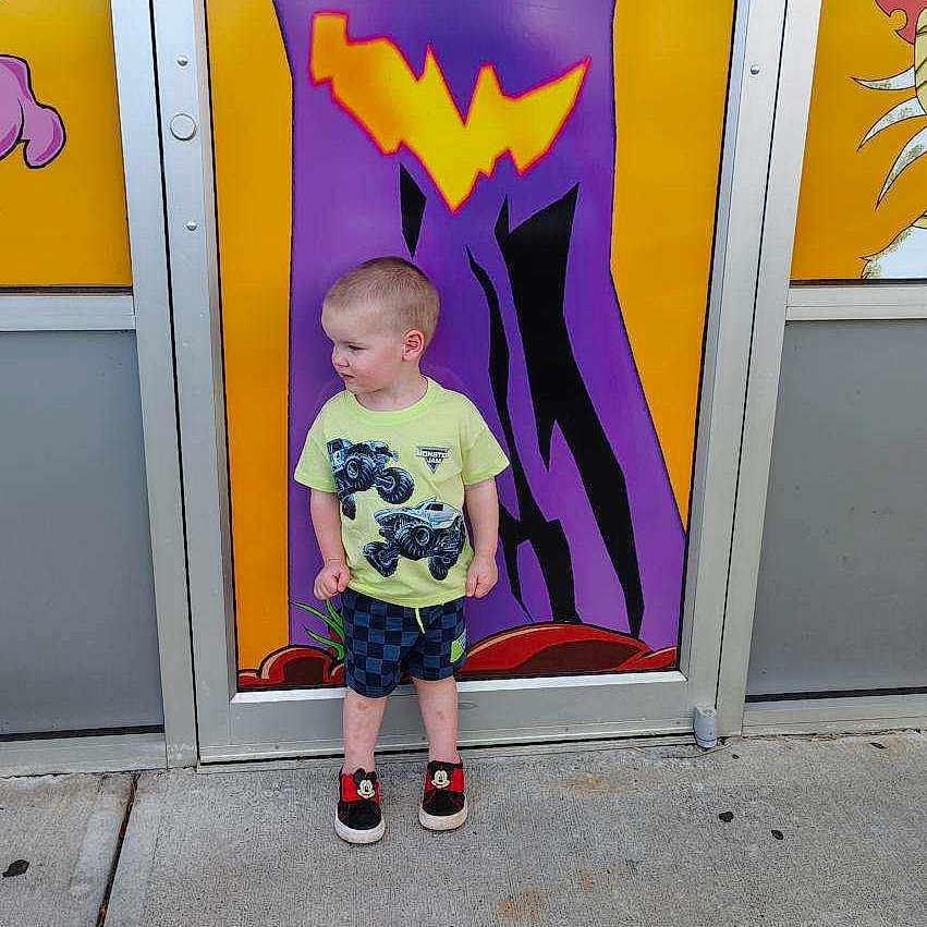 Joshua joined the competition — help win amazing prizes! child, boy, standing, sideways, yellow_shirt, monster_truck, checkered_shorts, mickey_mouse_shoes, concrete_sidewalk, colorful_background, halloween, jack_o_lantern, door, purple, orange, outdoor, curious_expression, casual_clothing, young_child, portrait