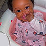baby, child, infant, smiling, sitting, portrait, curly_hair, dark_skin, pink_outfit, pajamas, cartoon_print, baby_seat, pacifier, finger_in_mouth, bright_eyes, indoor, wood_floor, cushion, happy, close_up