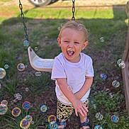 Rowan is registered to the contest to win money with this photo: toddler, child, outdoor, grass, swing, bubbles, happy, smiling, white_shirt, camouflage_shorts, sandals, sunlight, playground, nature, fun, joy, person, portrait, summer, daytime